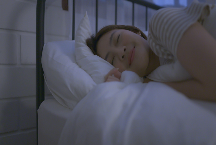 A woman sleeping soundly on a bed with white pillows and a striped shirt, captured in a cool-toned, softly lit bedroom setting.